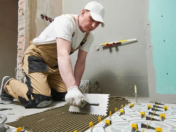 Worker installing floor tiles with a notched trowel in a room under renovation